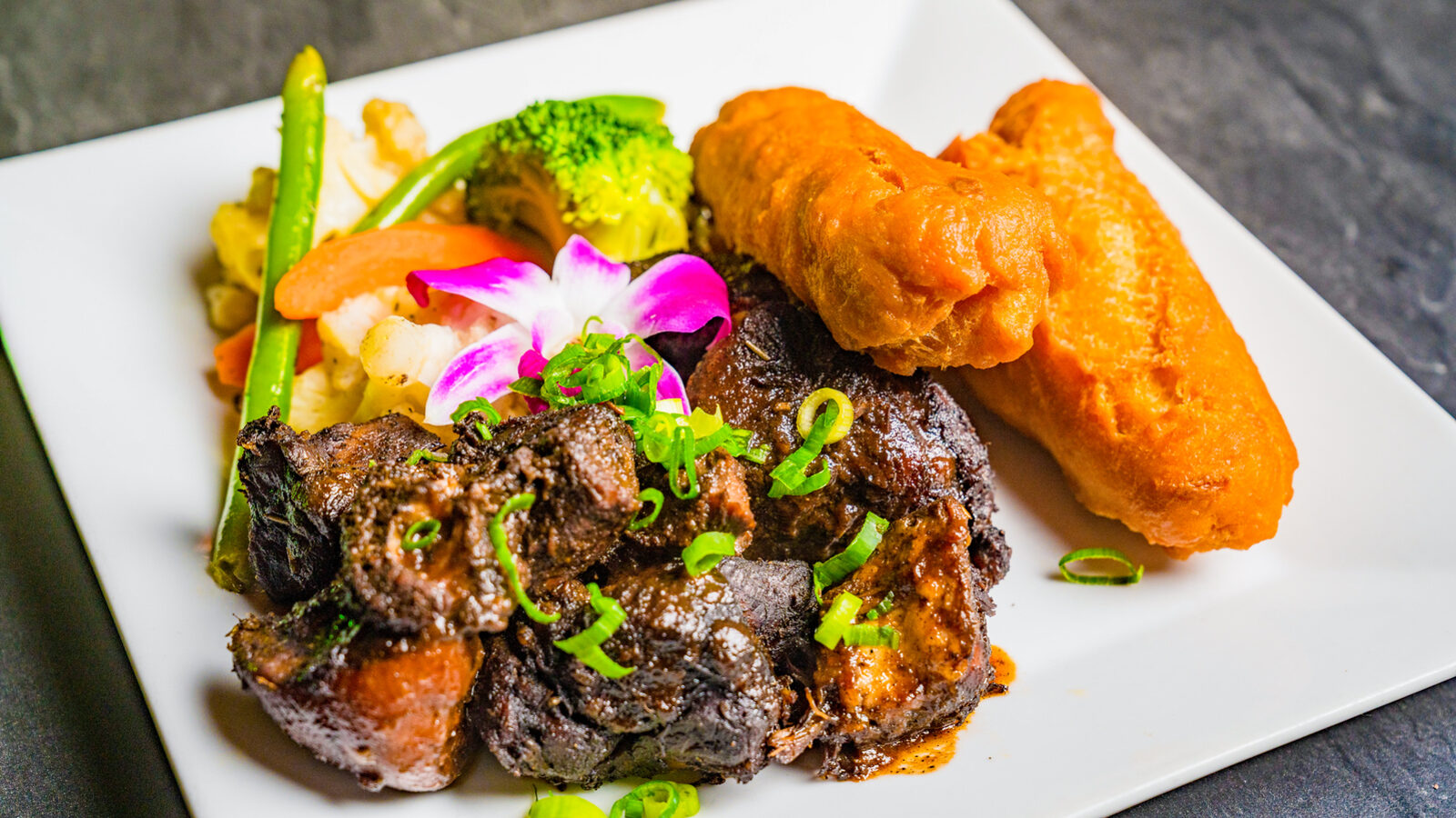 Braised oxtail with fried festival and vegetables on a white plate